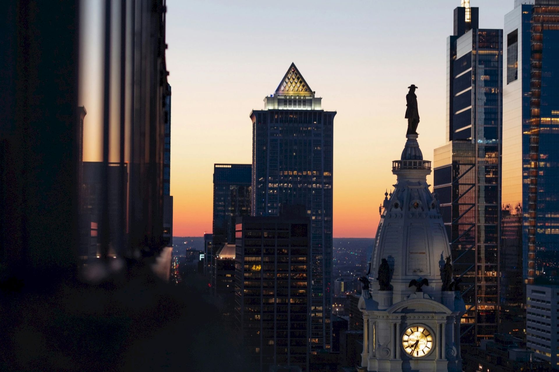 City skyline at sunset, featuring tall buildings and a clock tower with a statue on top, against a colorful sky.