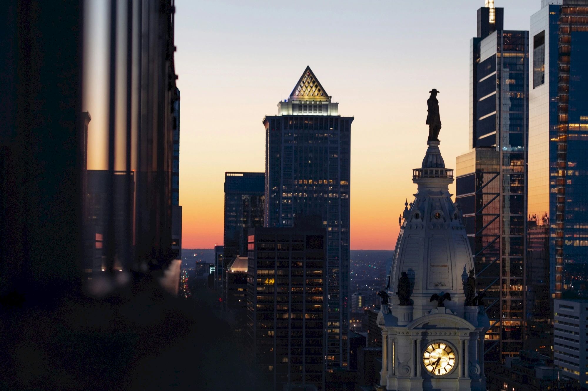 City skyline at sunset, featuring tall buildings and a clock tower with a statue on top, against a colorful sky.