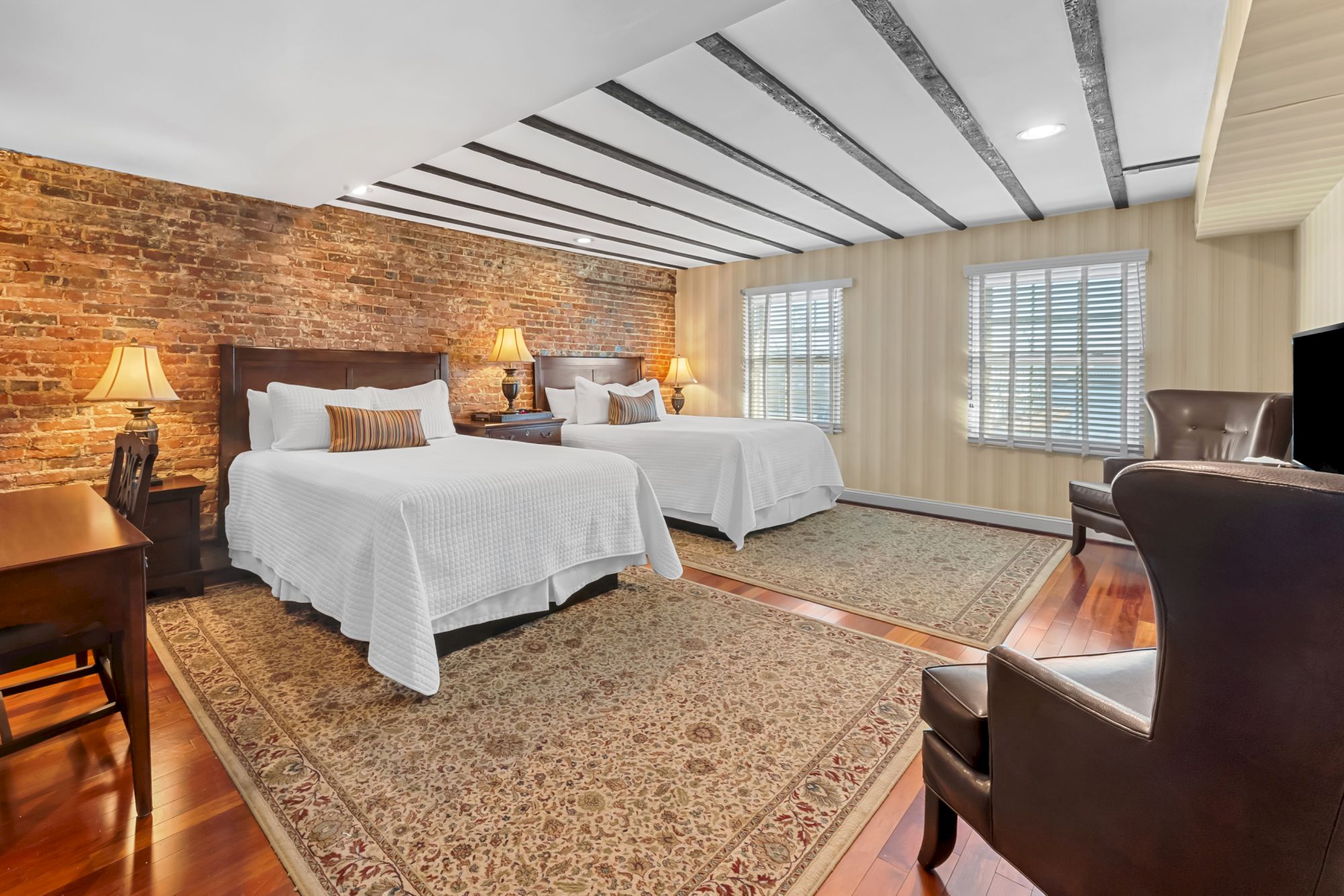 A cozy hotel room with two large beds, brick accent wall, warm lamps, wooden furniture, and a seating area by windows, bright and inviting.