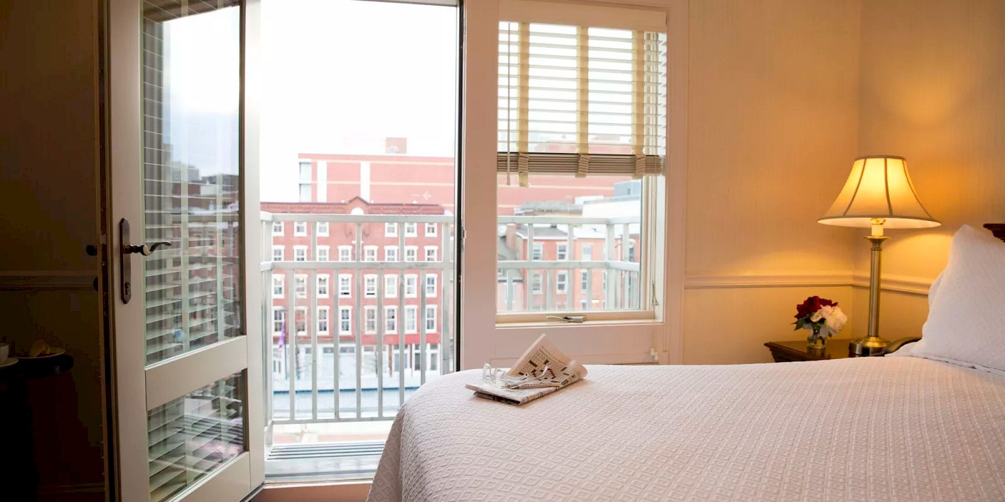 A cozy bedroom with a bed, a lamp on a nightstand, a balcony view, and an open book with glasses placed on the bed.