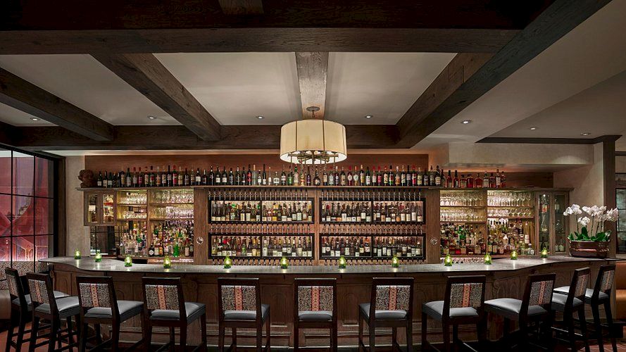 This image shows a well-stocked bar with rows of bottles, a long counter with stools, and a large overhead light fixture.