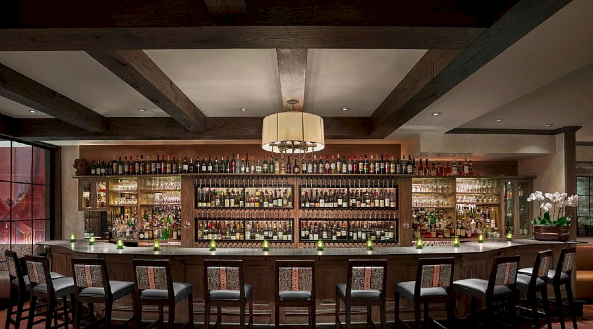 This image shows a well-stocked bar with rows of bottles, a long counter with stools, and a large overhead light fixture.