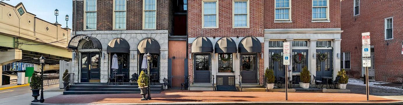 Historic brick buildings with arched doorways and tall windows along a sunny street, storefronts on the ground floor, blue sky above.