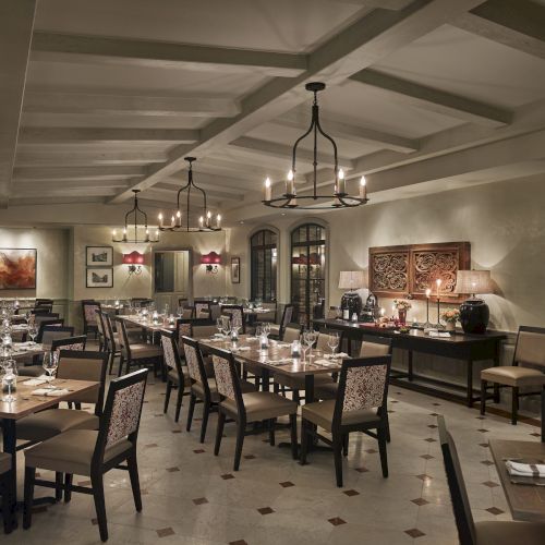 The image shows an elegantly set dining room in a restaurant with chandeliers, neatly arranged tables, and artwork on the walls.