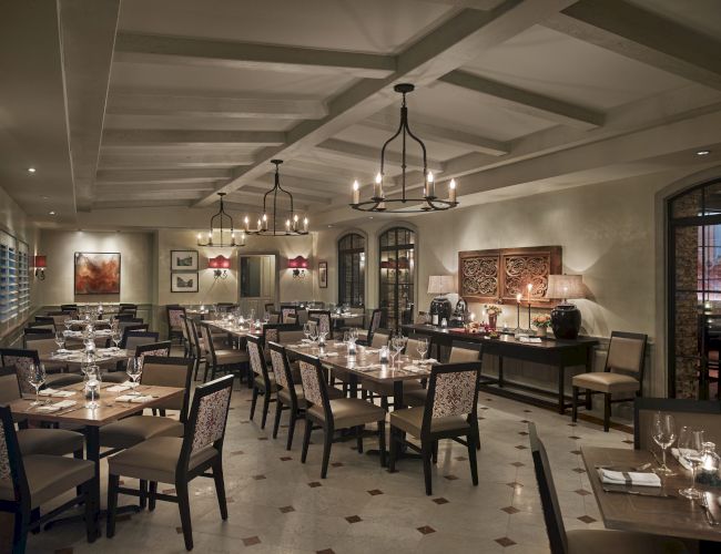 The image shows an elegantly set dining room in a restaurant with chandeliers, neatly arranged tables, and artwork on the walls.