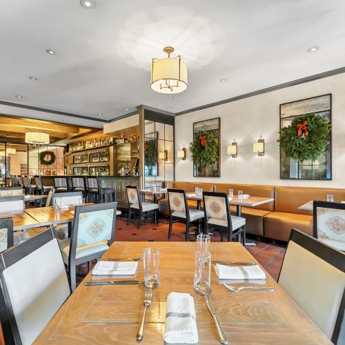 A bright, modern restaurant interior with wooden tables, cushioned booths, wall art, greenery, and an open bar area in the background.