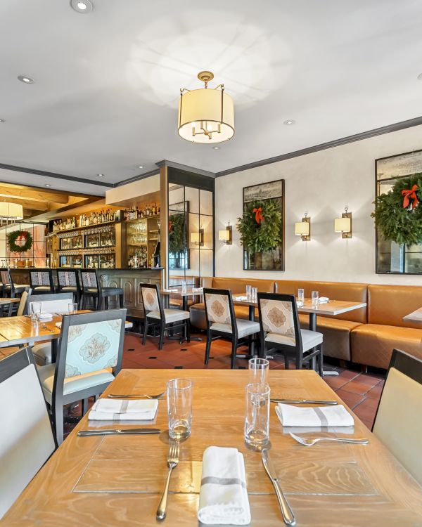 A bright, modern restaurant interior with wooden tables, cushioned booths, wall art, greenery, and an open bar area in the background.