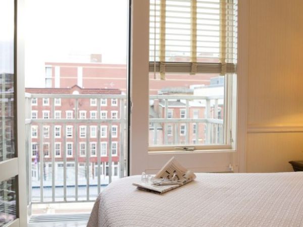 A cozy bedroom with a neatly made bed, a side lamp, flowers, and a view through glass doors showing a building outside.