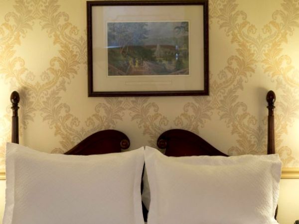 A cozy bedroom setup with two pillows, bedside lamps, a framed painting above the bed, and a small vase with roses on the left.