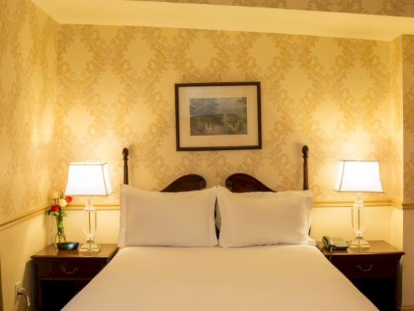 A cozy hotel room featuring a bed with two lamps, a framed picture on the wall, and elegant wallpaper.