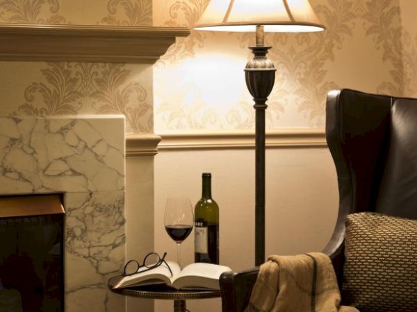 A cozy setting with a leather chair, a lamp, a marble fireplace, a book, glasses, a wine bottle, and a glass on a side table ends the scene.