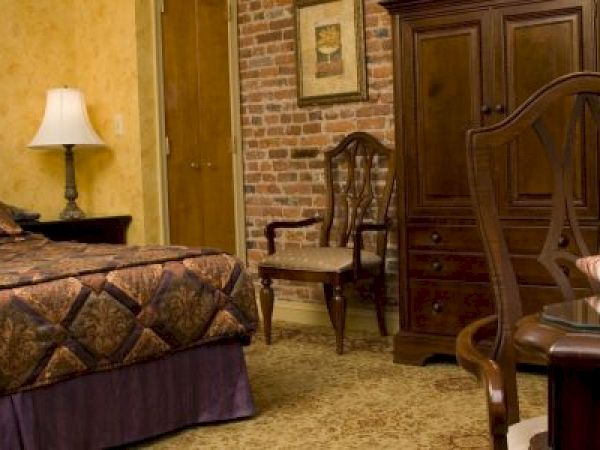 A cozy bedroom with a bed, lamp, wooden furniture, and exposed brick wall. A table holds a laptop, papers, and chair nearby.