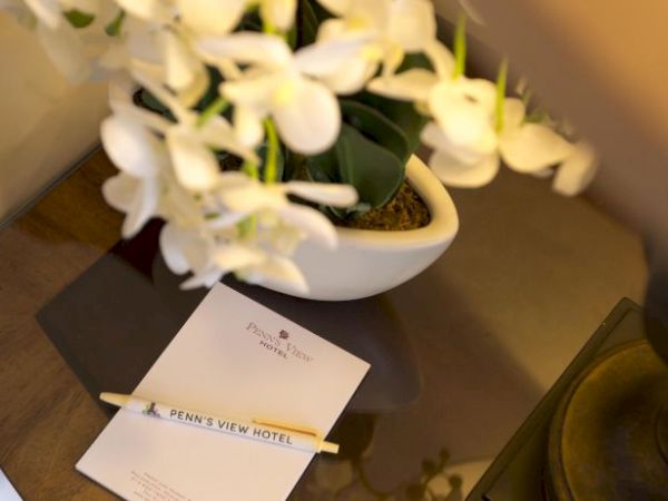 A bedside table with a lamp, notepad, pen labeled "Penn's View Hotel," and a vase of white flowers.