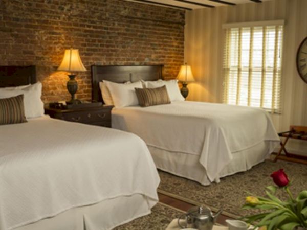 A cozy bedroom with two beds, brick wall, lamps, large clock, and flowers on a table, creating a warm and inviting atmosphere.