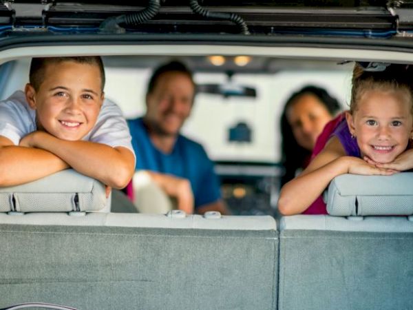A family is sitting in a car, with two children smiling at the back. The scene is cheerful and relaxed, suggesting a road trip.
