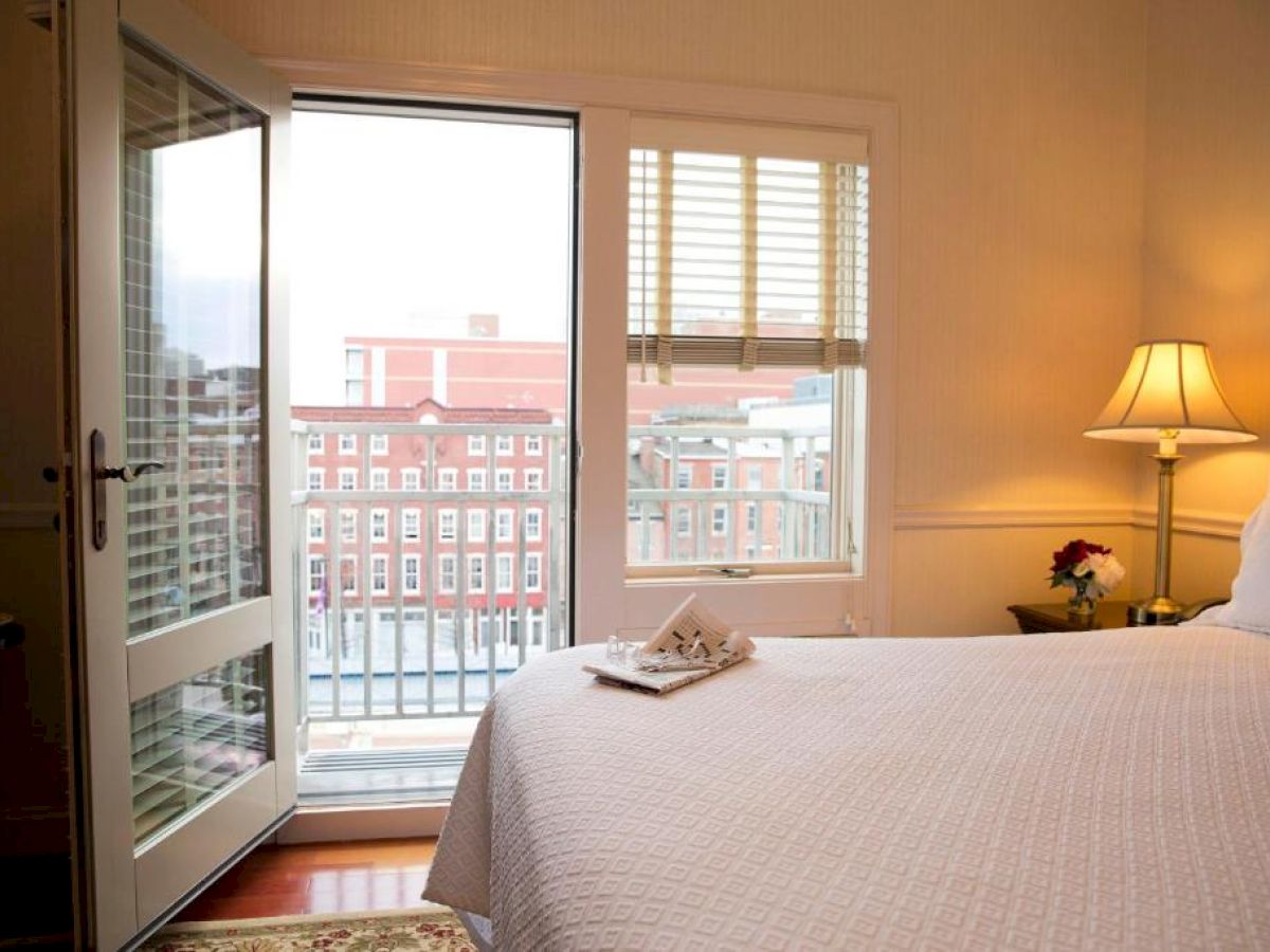 A cozy bedroom with a bed, nightstand, and lamp. A door opens to a balcony with views of red brick buildings outside.