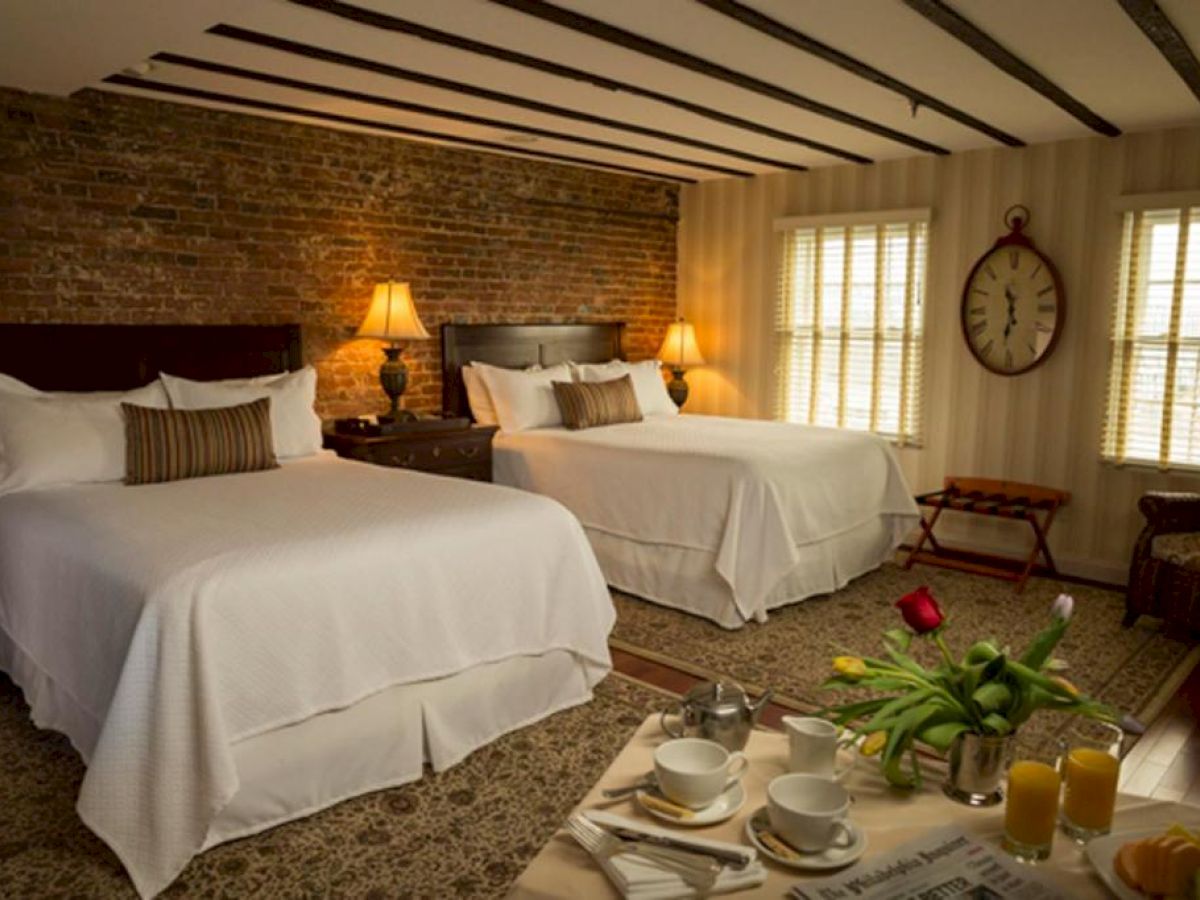 A cozy bedroom with two beds, a brick wall, lamps, a clock, and a table set with flowers and breakfast items.