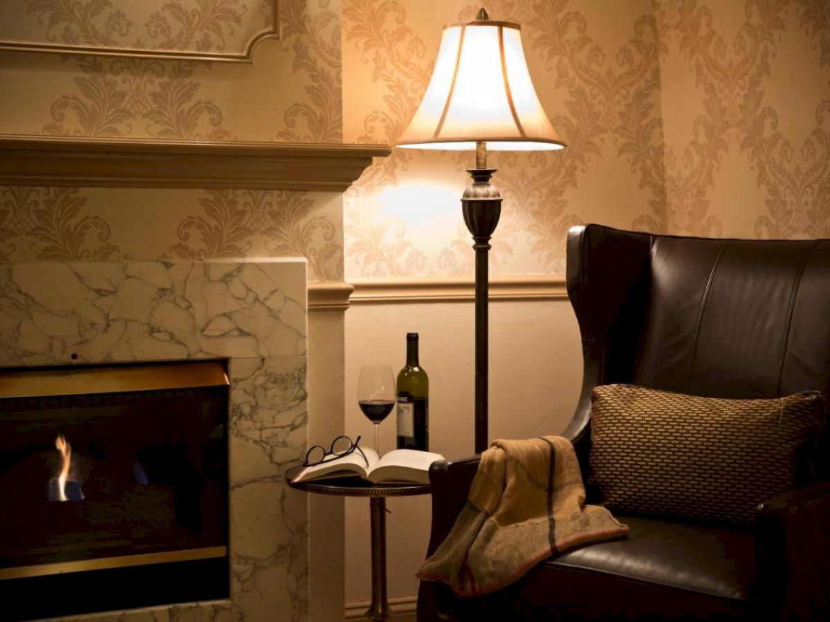 A cozy room features a leather chair, fireplace, lamp, open book with glasses, wine bottle, and glass on a side table.