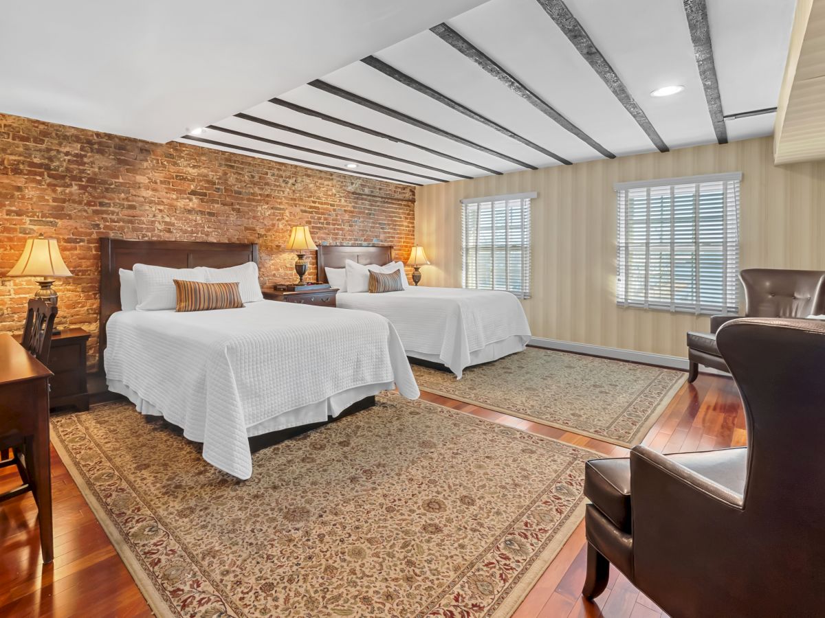 A spacious hotel room with two queen beds, exposed brick wall, matching lamps, a rug, windows with blinds, and a seating area with chairs.