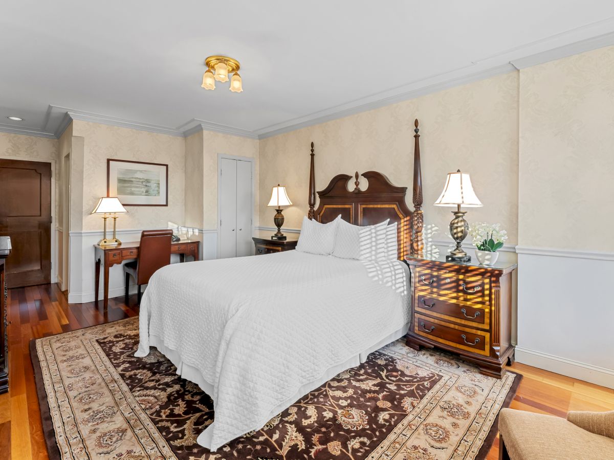 A luxurious hotel room with a large four-poster bed, matching nightstands, a patterned area rug, wooden furniture, and soft lighting.