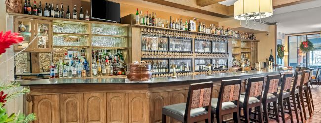 A cozy wood-paneled bar with a long counter, row of stools, and shelves full of bottles behind it, warm lighting, and tiled floor.