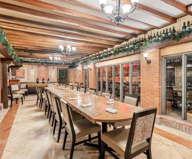 A long dining table with matching chairs runs down a wine-tiled room, flanked by glass-fronted wine storage, shelves of bottles, and warm overhead lighting, ready for a tasting.
