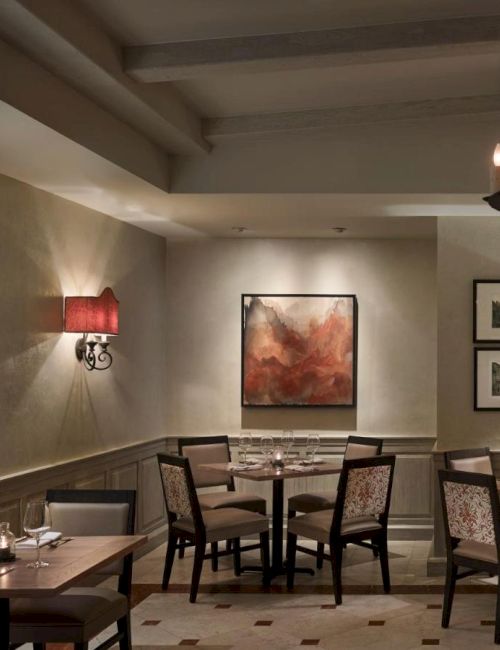 Cozy restaurant interior with neatly set tables, wall art, red-shaded sconces, and a candle-style chandelier, creating a warm ambiance.
