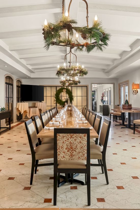 A long dining hall with a central table set for a formal meal, chandeliers, wreaths, and chairs along the sides, bright and orderly.