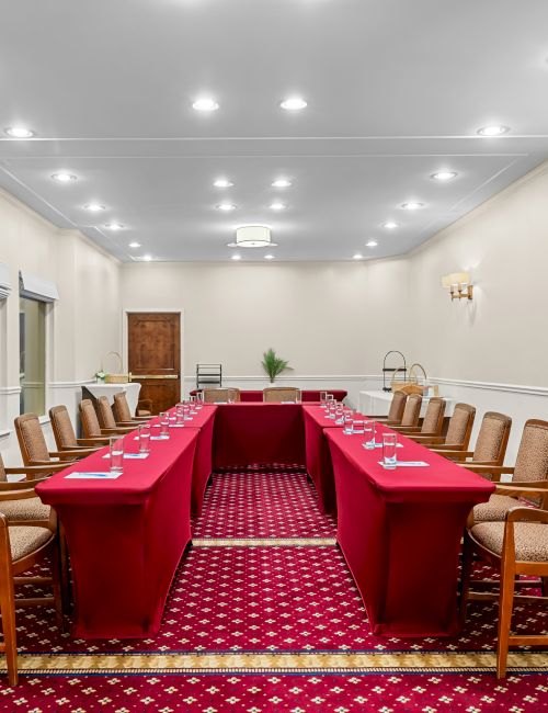 A long conference room with red-carpeted floor, two U-shaped tables, beige chairs, white walls, and a few wall lamps.