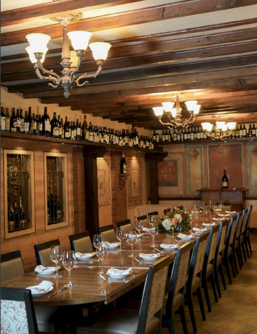 The image shows an elegant dining room with a long wooden table, set for a meal, featuring chandeliers and wine bottles on shelves.