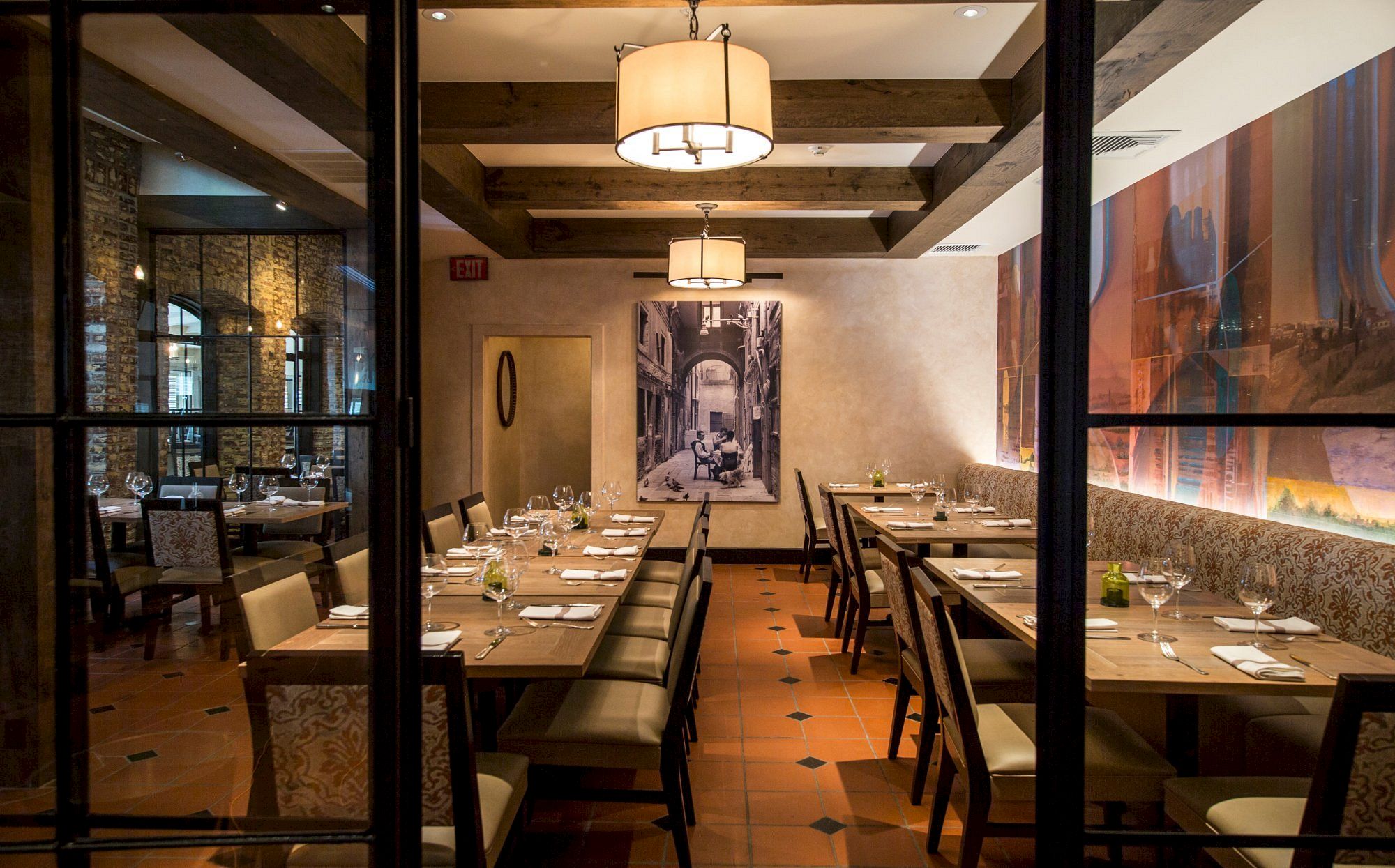 A warmly lit restaurant interior with wooden beams, elegant dining tables set with glasses and napkins, and artwork on the walls.