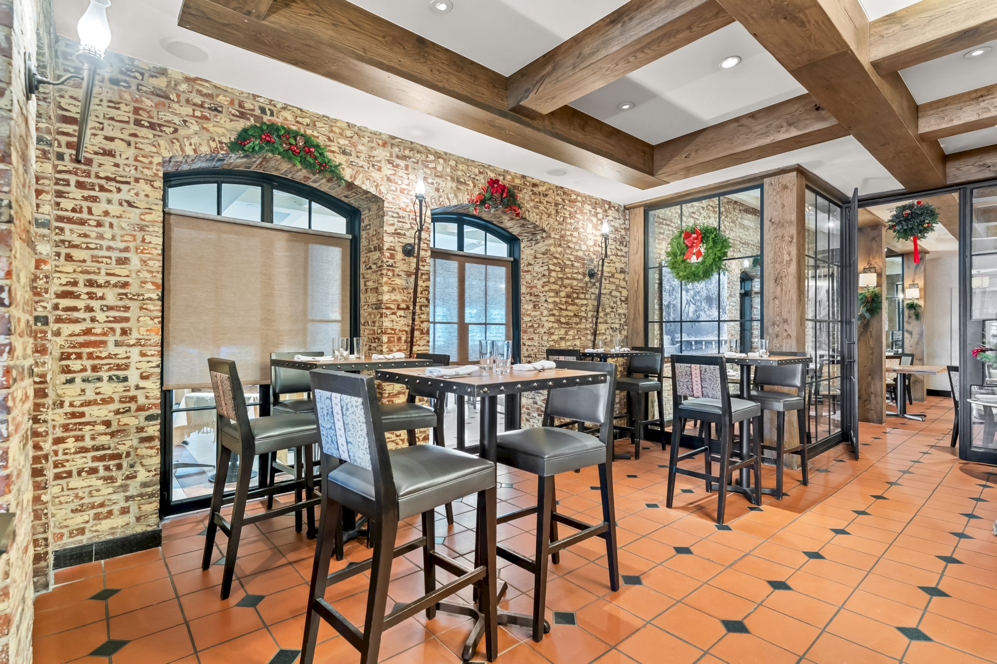 A stylish dining area with brick walls, arched windows, wooden ceiling beams, terracotta tiled floor, and multiple high tables with chairs, festive wreaths, and warm lighting.