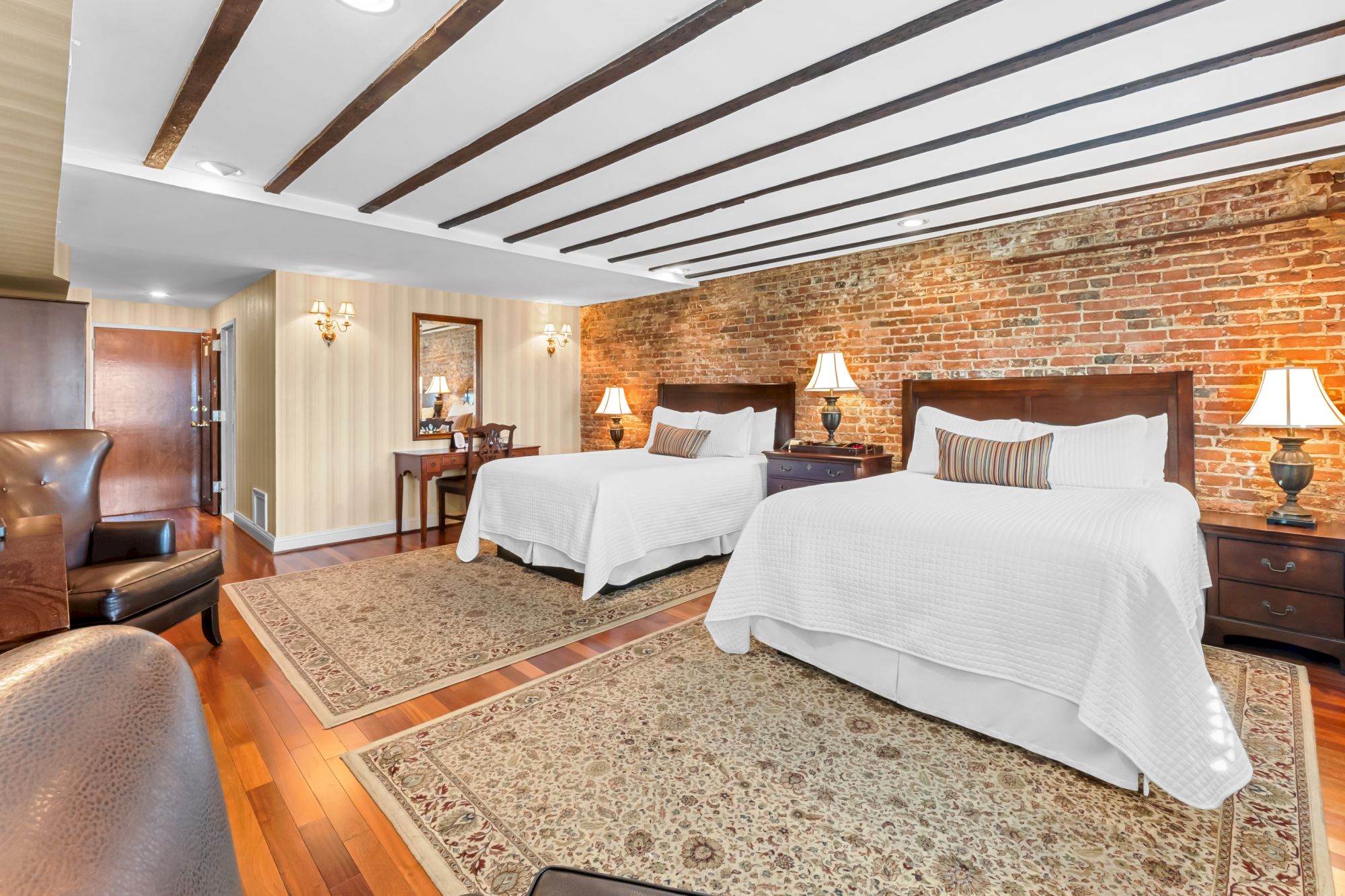 A hotel room with two large white beds, brick accent wall, wooden ceiling beams, lamps, and a patterned rug across polished hardwood floors.