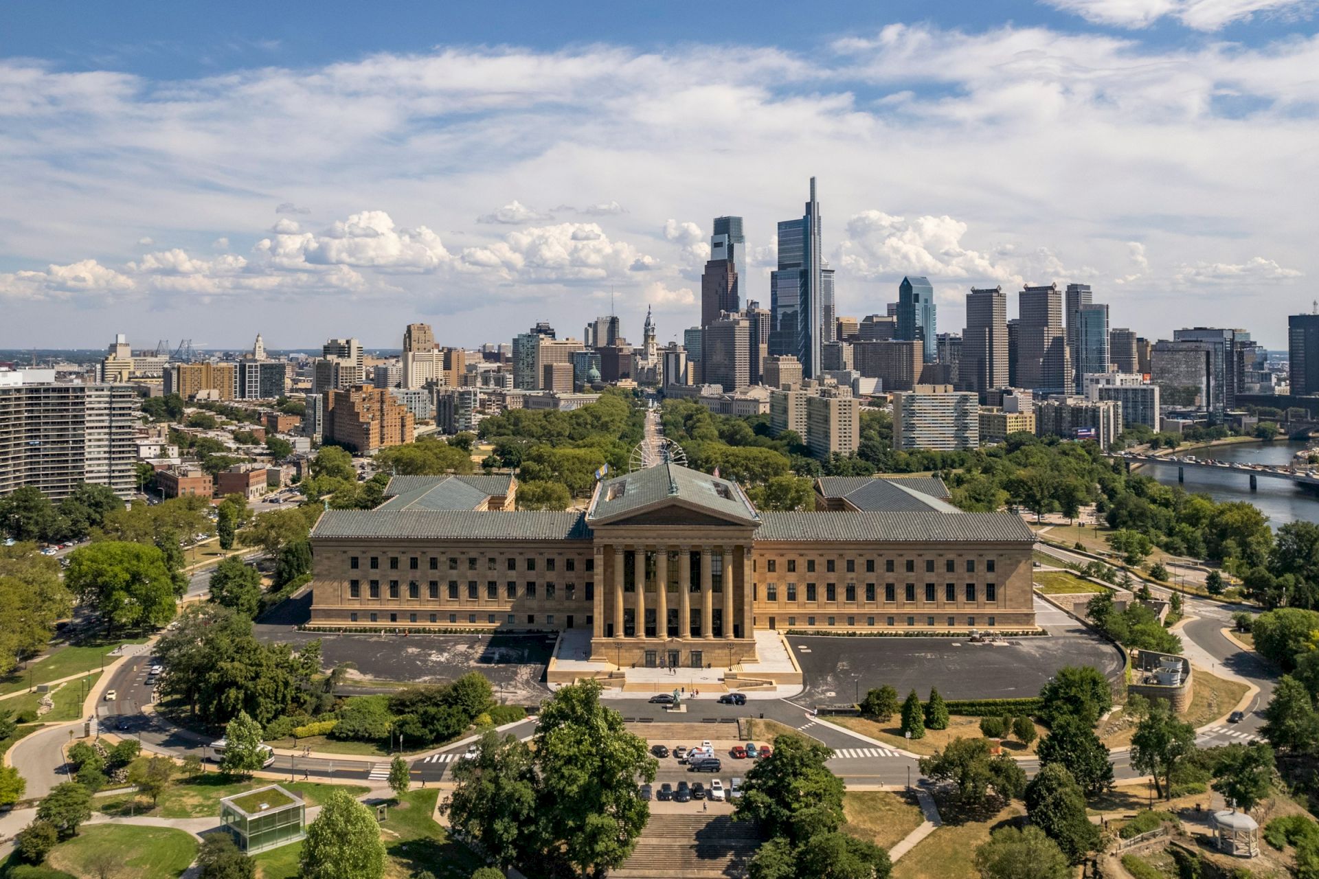 An aerial view of a large building with columns, surrounded by a cityscape and greenery, with a river visible on the right side.