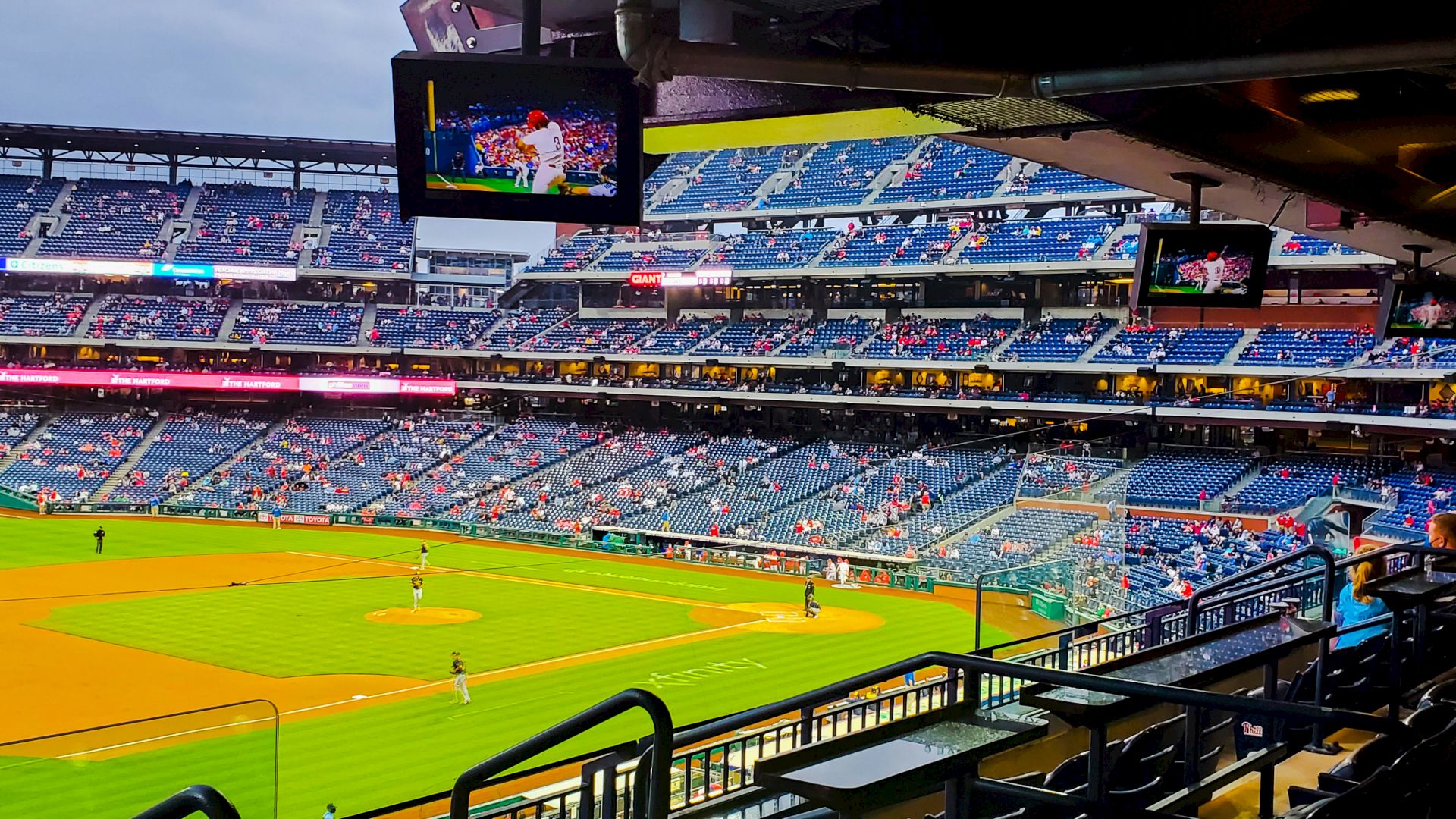 A baseball stadium with players on the field, seating areas partially filled, and screens showing the game.