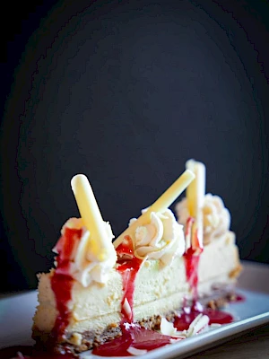 A slice of cheesecake with whipped cream, berry sauce, and white chocolate sticks on top.