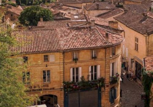 Aerial view of a charming European town with rustic buildings, red-tiled roofs, and cobblestone streets, surrounded by lush greenery.