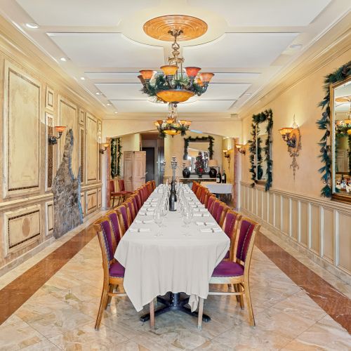 A long, elegant dining hall with a white-clothed table for many guests, gold accents, ornate chandeliers, and decorative mirrors along the walls.
