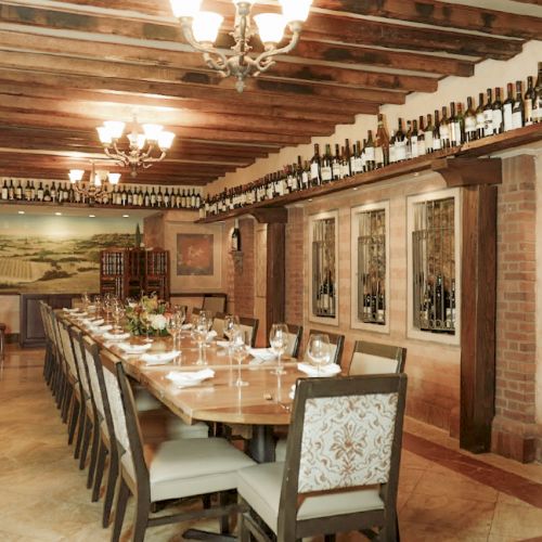 A long dining table in a rustic room with wine bottles on shelves, brick walls, chandeliers, and decorative art on the walls.