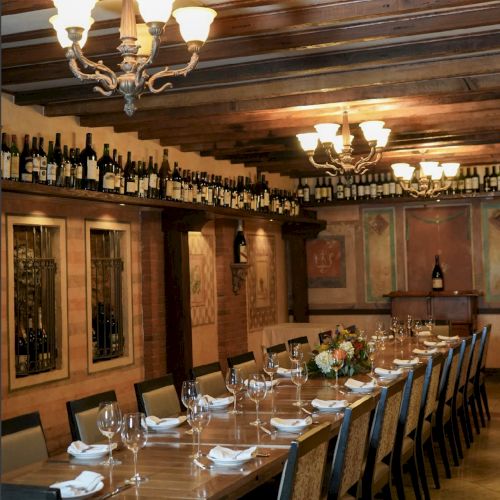 The image shows a long dining table set with wine glasses, napkins, and floral centerpieces in an elegant room with chandeliers and wine decor.