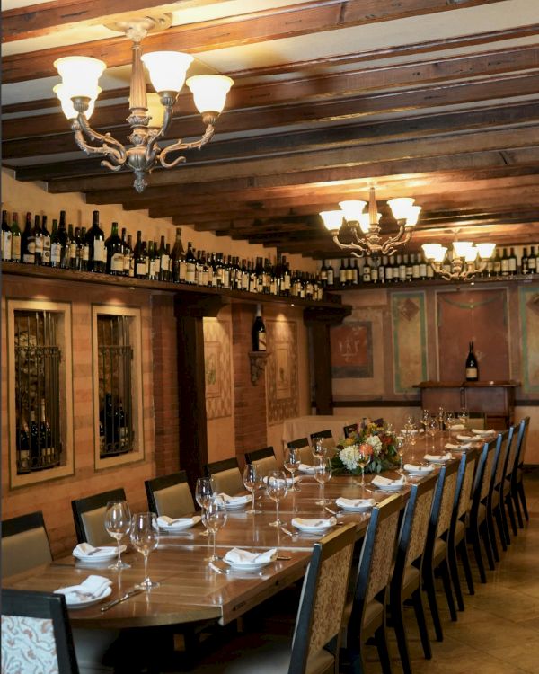 The image shows a long dining table set with wine glasses, napkins, and floral centerpieces in an elegant room with chandeliers and wine decor.