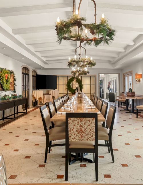 A long dining hall set for a formal meal, with a central table, upholstered chairs, wreaths, and decorative greenery along the room.