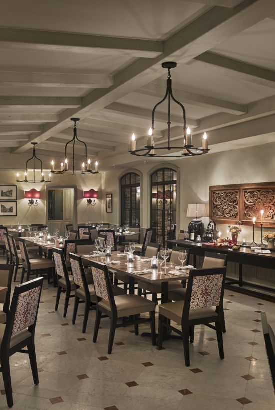 A cozy, elegant restaurant interior with neatly arranged tables, chairs, chandeliers, and warm lighting, creating an inviting atmosphere.