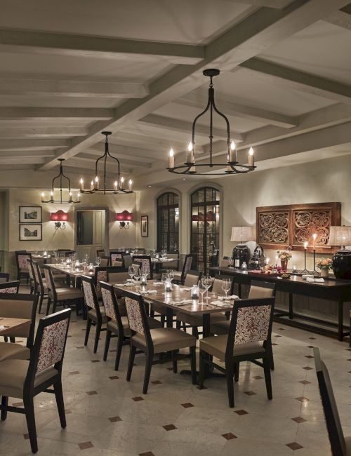 A cozy, elegant restaurant interior with neatly arranged tables, chairs, chandeliers, and decorative wall art, creating an inviting ambiance.