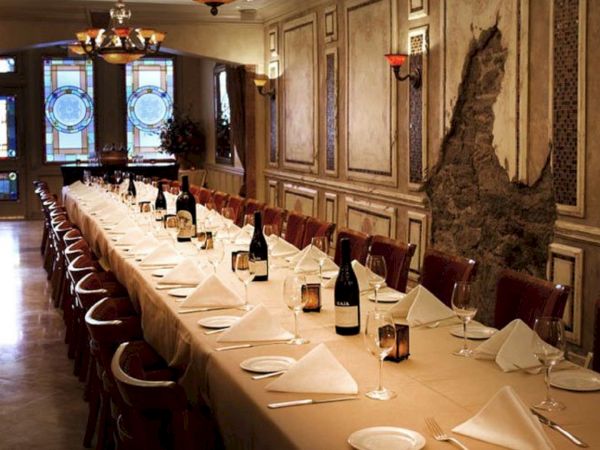 An elegant dining room with a long, set table, adorned with wine bottles and glasses, under warm lighting and stained glass windows.