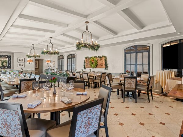 A bright, elegant dining room with round tables, upholstered chairs, chandeliers, and a buffet area in the back, ready for a formal meal.