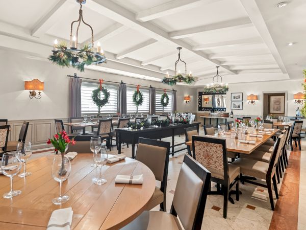 A bright, elegant dining room with long tables set for a formal meal, chandeliers, wreaths, and place settings ready for guests.