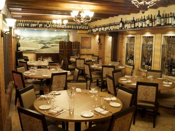 A cozy restaurant interior with wooden tables, chairs, and wine bottles lining the walls, under warm lighting and decorative ceiling beams.