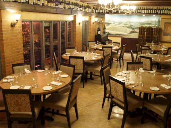The image shows a cozy restaurant interior with round tables set for dining, wine storage, chandeliers, and a mural on the wall.