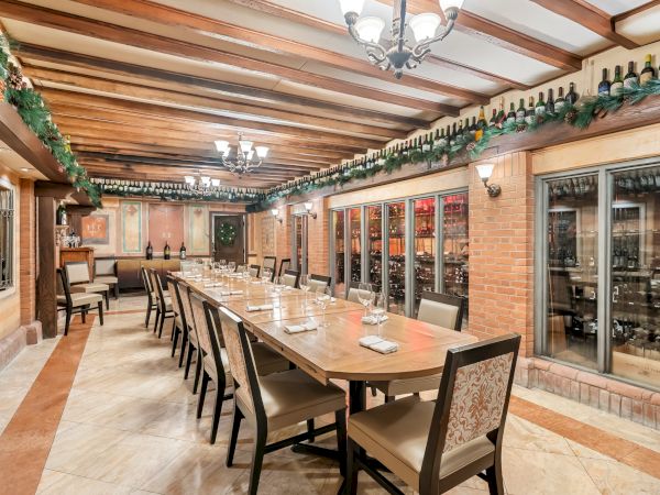 A long dining table with chairs in a wine cellar-like room, bottles on shelves along the walls, warm lighting, and decorative ceiling beams, ending the sentence.